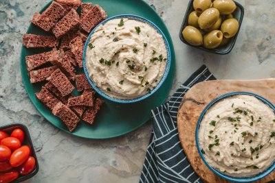 Two different bowls of butter bean dip served with bread olives and tomatoes on the side