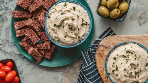 Two different bowls of butter bean dip served with bread olives and tomatoes on the side